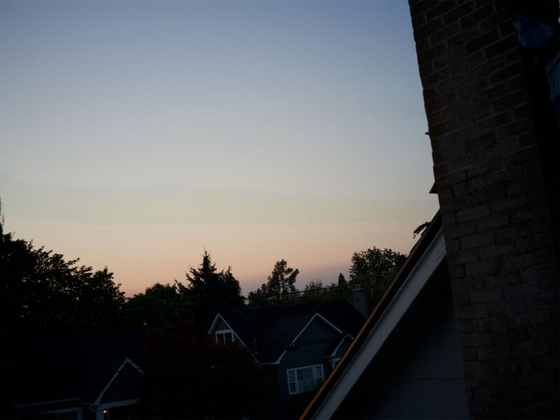 A sunset view over suburban rooftops with a clear sky transitioning from blue to soft pink and orange hues. Trees and houses are silhouetted in the dimming light, creating a peaceful evening scene.