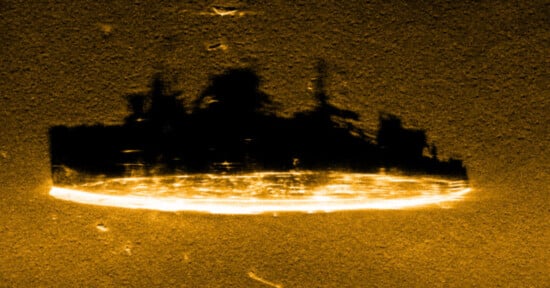 A sonar image shows a large, disk-shaped object resting on the seabed. Its rough, curved silhouette has bright reflections outlining its perimeter, resembling a UFO. The background appears grainy and dark.