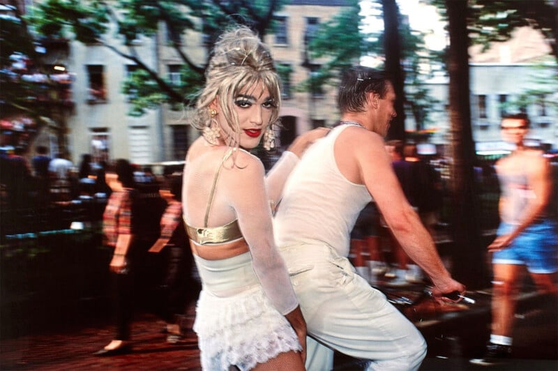 A person in a white outfit with a lacy skirt looks over their shoulder on a bustling street. They have dramatic makeup and a blonde updo. Nearby, a person wearing white rides a bicycle. The background features blurred pedestrians and trees.