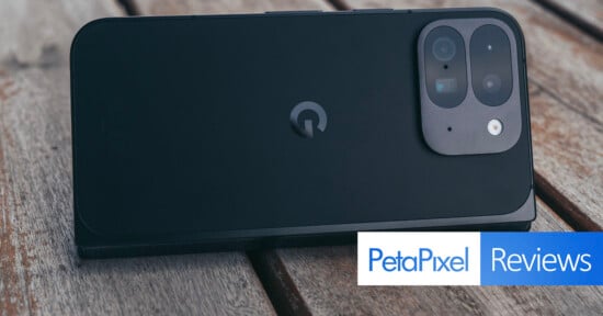 A smartphone with a dual-camera setup is placed on a wooden surface. The phone is black with a distinctive logo in the center. The bottom right corner has a "PetaPixel Reviews" logo.