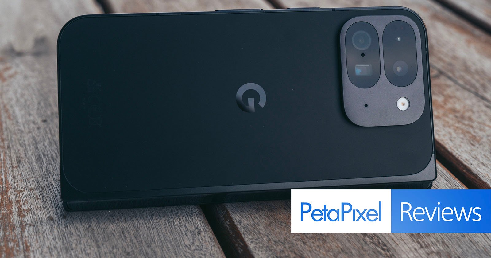 A smartphone with a dual-camera setup is placed on a wooden surface. The phone is black with a distinctive logo in the center. The bottom right corner has a "PetaPixel Reviews" logo.