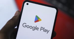 A person holds a smartphone displaying the Google Play logo, featuring a multicolored play button and the words "Google Play" on a white screen. The background is blurred, focusing on the phone's screen.