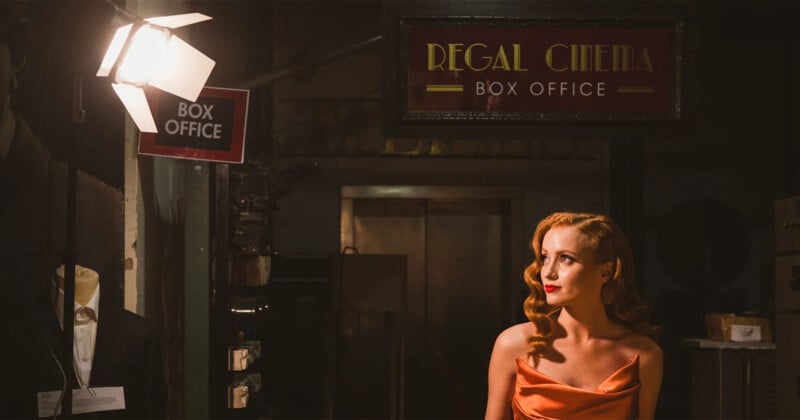 A woman with long, wavy hair in an elegant, sleeveless orange dress stands illuminated near a "Regal Cinema Box Office" sign. She looks off to the side, with soft, warm lighting creating a vintage cinematic atmosphere.