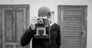 A person wearing a knit cap holds an old-fashioned camera up to their face, as if taking a picture. They stand in front of two wooden doors in this black and white image.