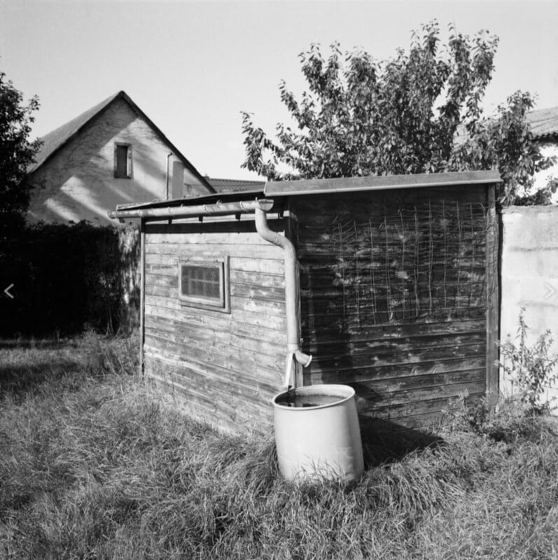 A black and white image of a small wooden shed with a corrugated metal roof, featuring a rainwater barrel connected to a downspout. Overgrown grass surrounds the shed, and a tree and house are visible in the background.