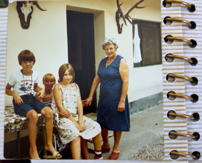 A vintage photo of four people outside a house. An older woman stands smiling beside two children seated on a bench, with an older seated man partially visible. Animal skulls with antlers adorn the wall behind them.