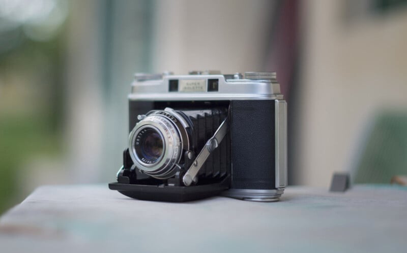 A vintage folding camera with a textured black leather and silver body is placed on a wooden surface. The lens is extended, and the background is softly blurred, suggesting an outdoor setting.