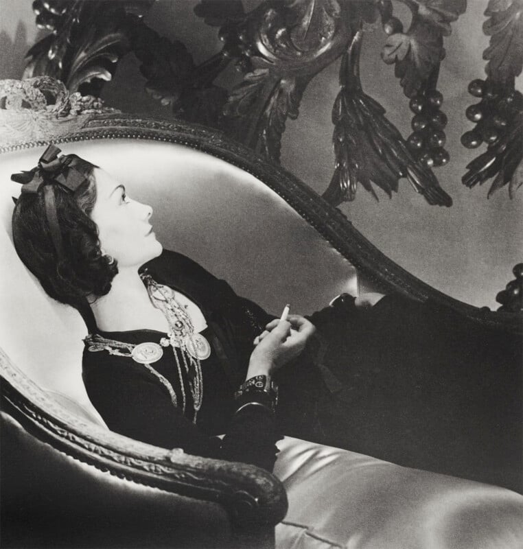 A black and white photo of a person in elegant attire lying on a luxurious, ornate chaise lounge. They are gazing sideways, wearing a dark outfit with layered necklaces, and are set against a richly decorated backdrop.