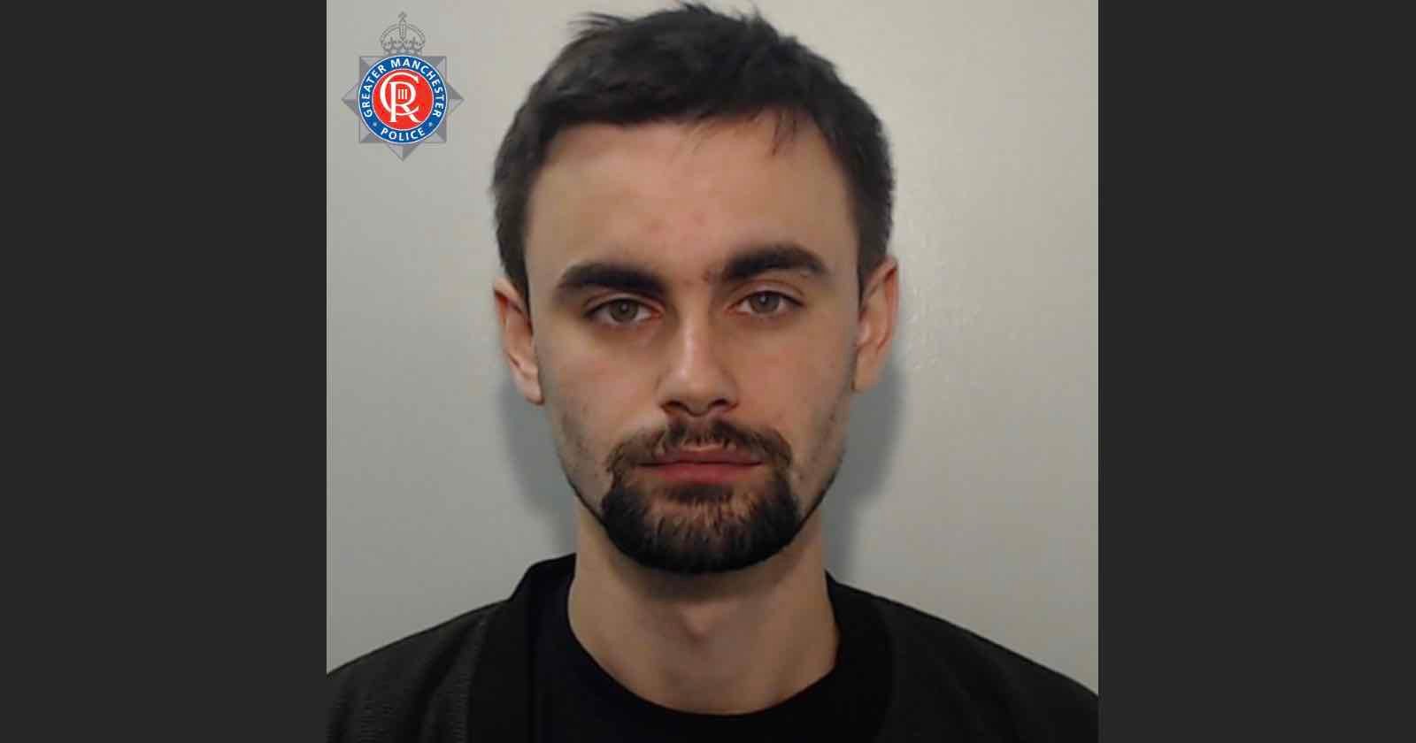 A man with short dark hair and a beard is shown in a mugshot. He wears a dark shirt, and there is a police insignia in the upper left corner. The background is plain and light-colored.