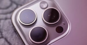 Close-up of a smartphone's rear camera system featuring three lenses and a flash, set against a blurred gray background. The lenses are arranged in a triangular pattern.