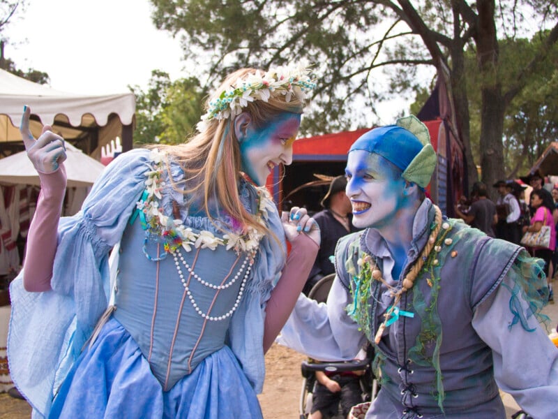 Two people in blue costumes and face paint, resembling aquatic-themed characters, smile at each other at an outdoor festival. One wears a flower crown, the other a blue cap. Tents and a crowd are visible in the background.