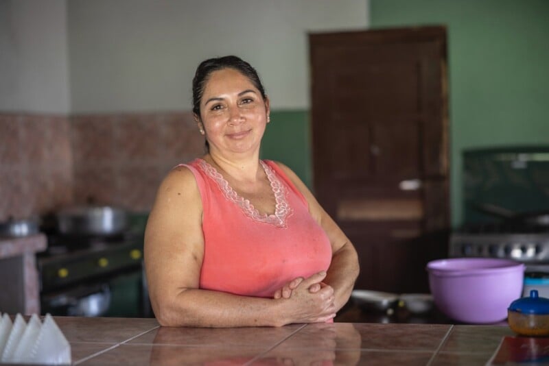 A woman in a pink sleeveless top smiles warmly while standing in a kitchen. She rests her arms on a tiled countertop. In the background, there are kitchen appliances and a purple bowl. The setting has an inviting, homely atmosphere.