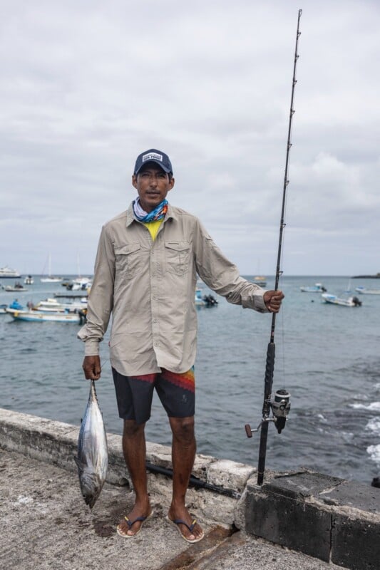 A man stands on a pier holding a fishing rod and a fish. He is wearing a beige shirt, colorful shorts, and a cap. The ocean and several boats are visible in the background under a cloudy sky.