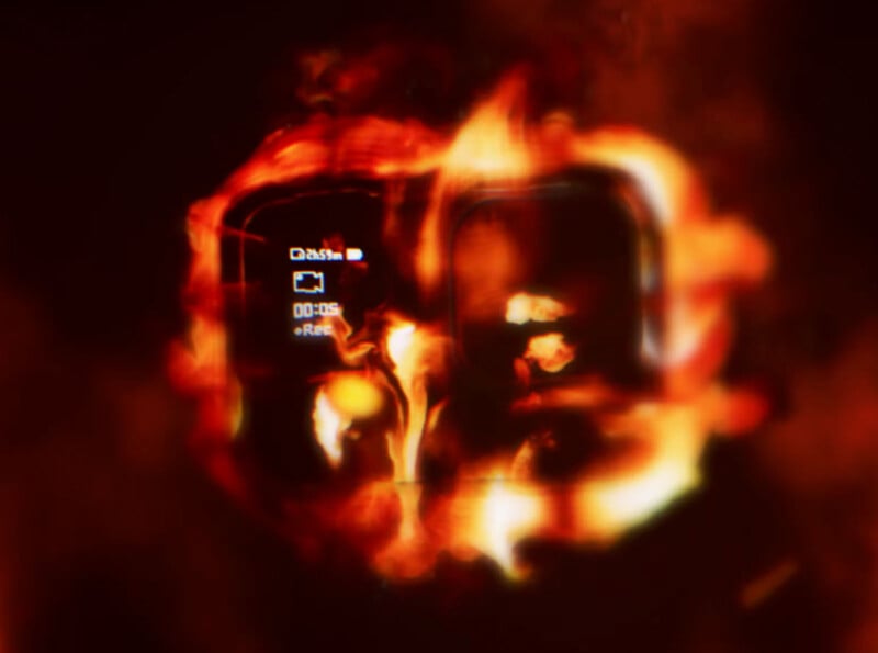 A digital camera with a small display screen and buttons is engulfed in bright, swirling flames, highlighting a contrast between technology and fire. The numbers "00:05" and "Rec" are visible on the screen.