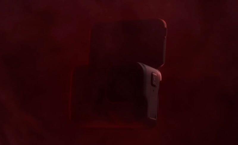 A dark, partially obscured image showing a boxy object with a subtle red hue. It appears to be a compact, rectangular shape with rounded edges, possibly resembling a camera or similar device. The lighting is dim, enhancing the mysterious atmosphere.