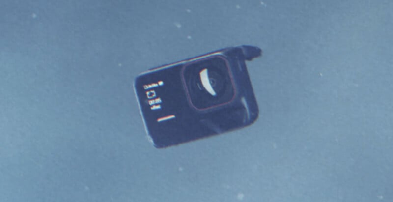 A black action camera is submerged underwater, tilted at an angle. The camera lens and a digital display screen showing basic settings are visible. The background is a bluish underwater scene.