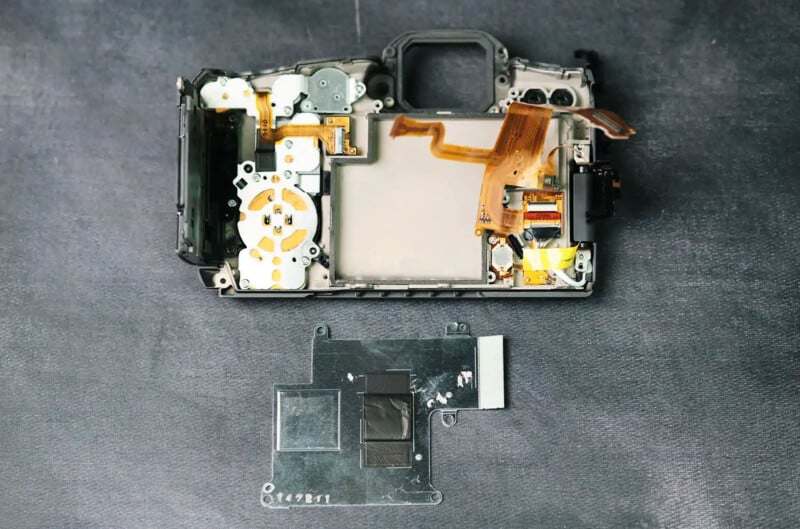 A disassembled camera lies on a dark surface, showing its internal components. The empty black frame and intricate circuits are visible, along with a detached piece placed beneath, revealing mechanical parts and wiring.