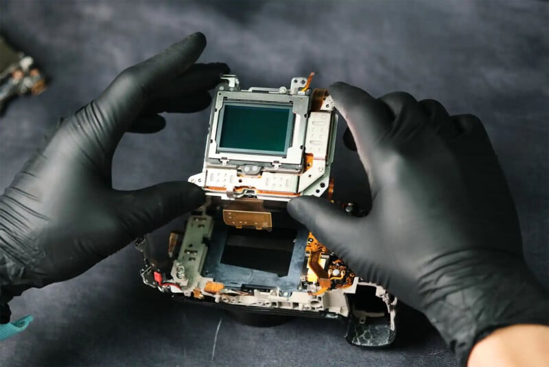 Hands in black gloves are delicately assembling or repairing a camera sensor. The image shows the internal components of a camera being worked on, emphasizing precision and care in handling delicate technology.