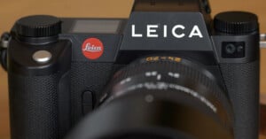 Close-up of a Leica camera displaying the brand logo prominently. The camera has a textured grip and a large lens attached, with the Leica emblem in red on the body. The image highlights the camera's design and brand identity.