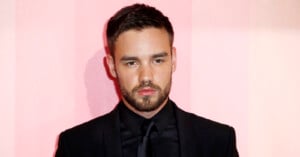 A man with short dark hair and a beard is dressed in a black suit and tie, standing against a light pink backdrop.