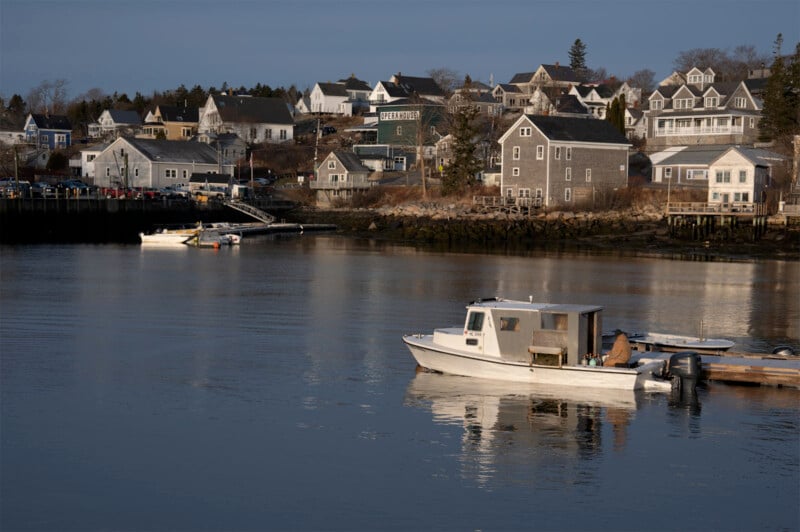 A serene coastal village with wooden houses on a hillside, overlooking a calm harbor. A small white boat is docked on the water, with early evening light casting a peaceful ambiance.