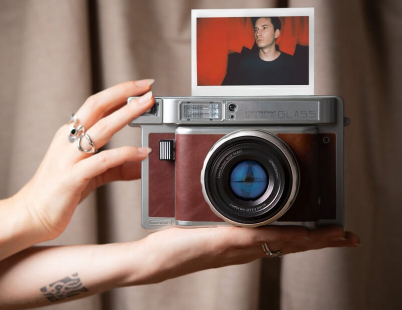 A person holding a vintage-style instant camera with a developing photo on top, showing a person in a red room. The camera features a large lens and a leather body. The holder has a tattoo and wears multiple rings.