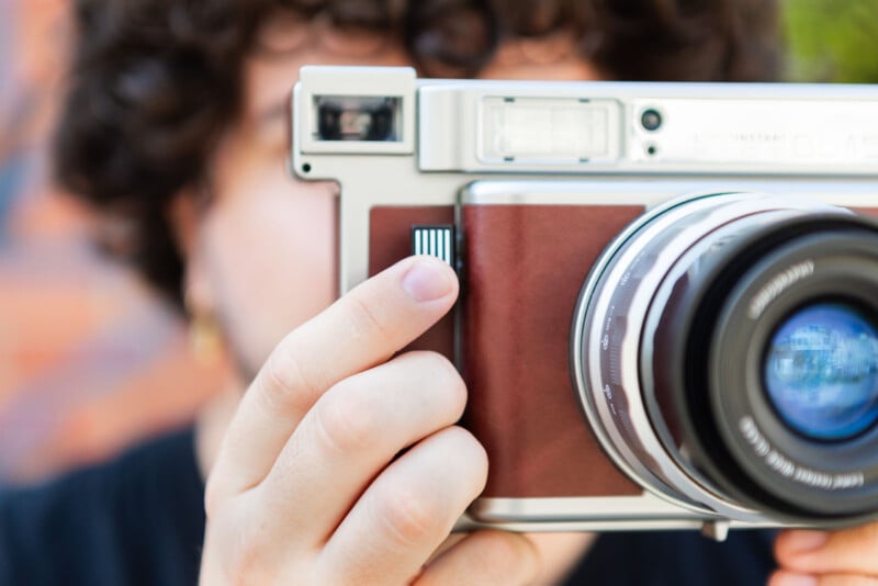 A person with curly hair is holding a vintage-style camera close to their face, positioning their fingers on the side of the camera. The camera lens is prominently visible in the foreground.