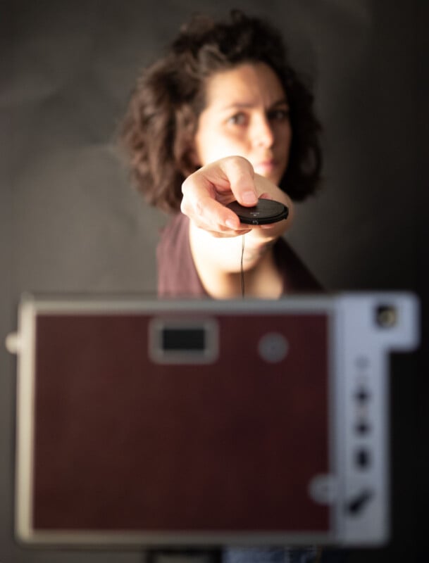 A person with curly hair is holding a camera remote control, pointing it toward the camera. The focus is on the remote, and a blurred camera lens is visible in the foreground. The background is dark and out of focus.