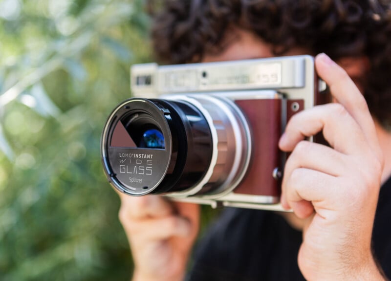 A person with curly hair holds a large vintage-style instant camera close to their face, preparing to take a photo outdoors. The camera is labeled "Lomoinstant Wide Glass Splitzer," and blurred greenery is visible in the background.