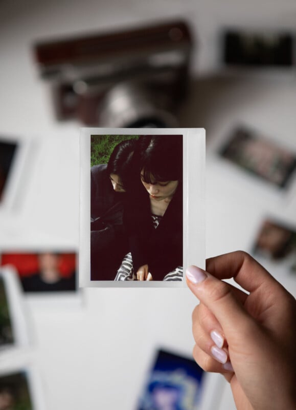 A hand holds a Polaroid photo of two people sitting closely, with their heads slightly bowed. Around the table, there are other blurry photos and an old camera in the background.