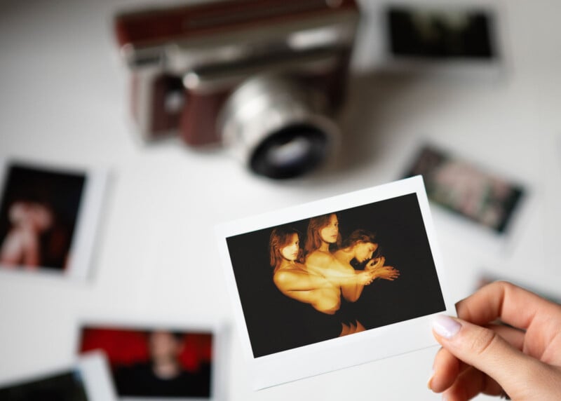 A hand holds a developed photo showing three people posing against a black background. The photo is surrounded by other blurred images and a vintage camera on a white surface.