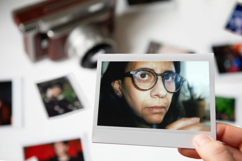 A person holds a glossy photograph showing a close-up of someone wearing large glasses. In the background, an old-fashioned camera and scattered photos can be seen on a white surface.