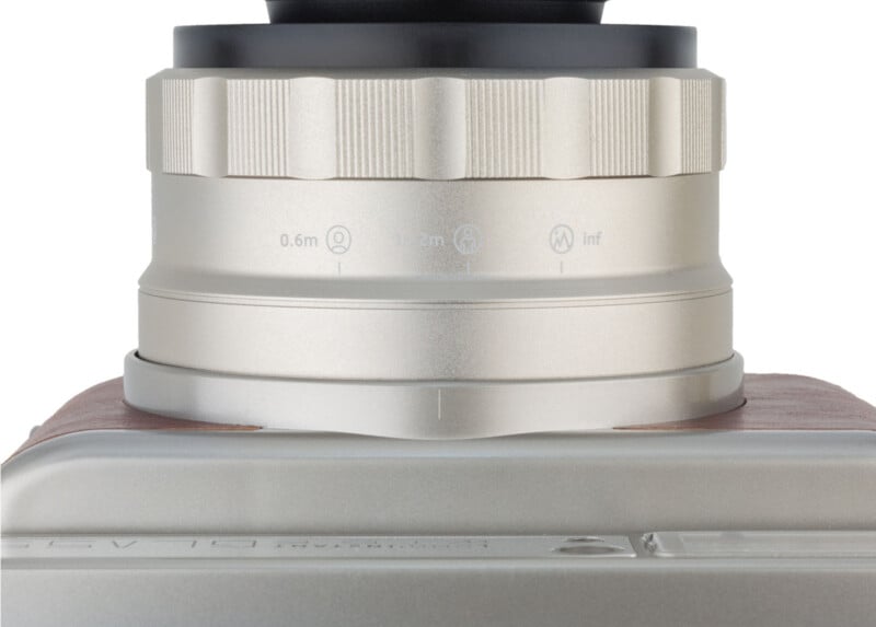 Close-up of a camera's silver lens and body, showing focus markings and textured surfaces. The lens is partially extended, revealing details like distance indicators. The camera body features a sleek, metallic finish.