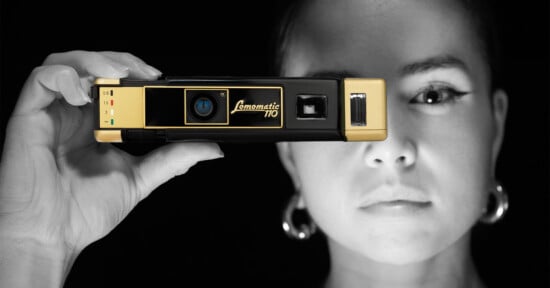 A person holds a vintage Lomomatic 110 camera up to their eye, partially obscuring their face. The camera is black and gold, and the background is dark. The person wears large hoop earrings and the image is in black and white except for the camera.