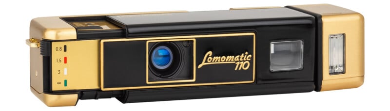 A vintage black and gold Lomomatic 110 camera with a blue lens. The design features labeled settings on the left, a prominent logo in the center, and a flash module on the right.