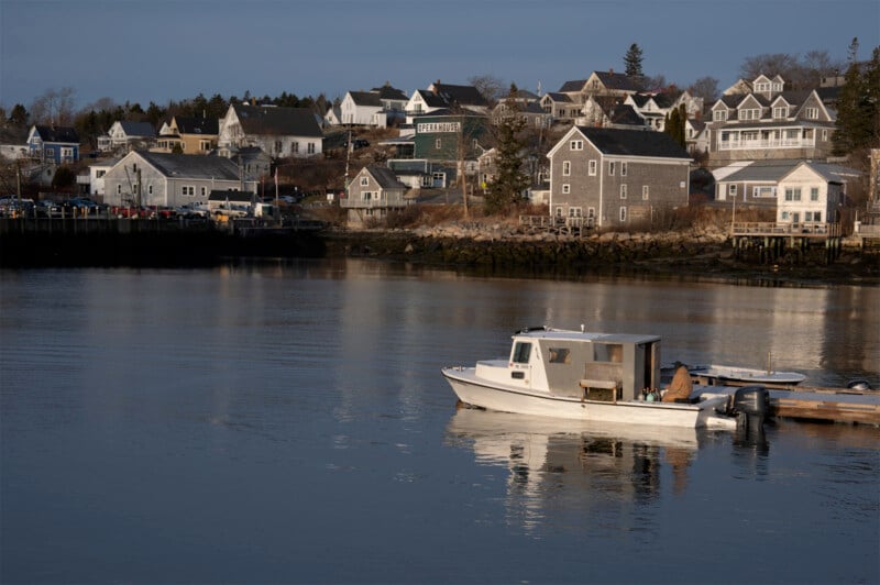 A serene coastal scene featuring a quiet harbor with a docked white fishing boat. Houses with weathered wood siding line the hillside in the background, under a clear blue sky. The calm water reflects the buildings and boat, creating a peaceful atmosphere.