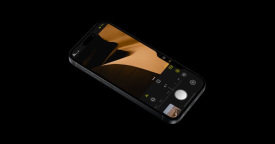 A smartphone with a sleek black frame is displayed against a black background. The screen shows a photography interface with an image of sand dunes. The interface includes various camera settings and icons, with a large shutter button at the bottom.