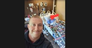 A man takes a selfie in a hospital room. He smiles in the foreground, while a person rests in a hospital bed in the background, covered with a colorful quilt. Medical equipment surrounds the bed.