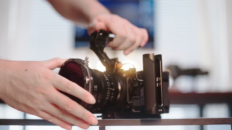 Person adjusting a professional video camera on a tripod in a well-lit room. The camera has a large lens and various attachments.