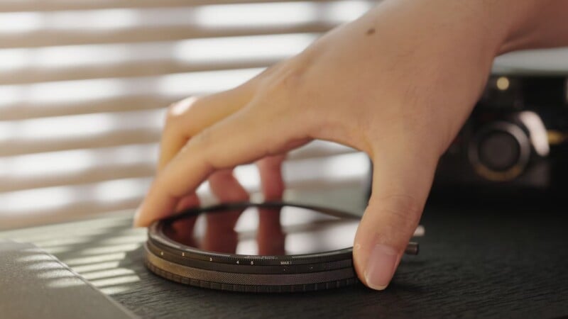 A hand adjusting a stack of camera lens filters on a table, with window blinds casting soft sunlight and shadows in the background.