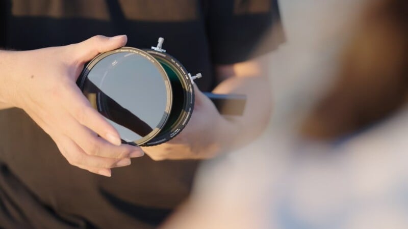 A person holds a neutral density filter for a camera with both hands. The filter is round and dark, set against a blurred background. The person wears a black shirt, and the scene is lit by natural light.