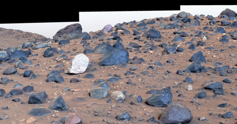 A rocky Martian landscape with various sizes and shapes of blue-tinted boulders scattered across a reddish-brown sandy terrain. The ground slightly rises in the background under a pale sky.