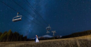 A couple embraces under a starry night sky, with ski lift chairs suspended overhead. The horizon shows a snow-capped mountain and a forest, creating a romantic and serene atmosphere.