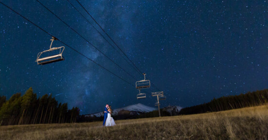 A couple embraces under a starry night sky, with ski lift chairs suspended overhead. The horizon shows a snow-capped mountain and a forest, creating a romantic and serene atmosphere.