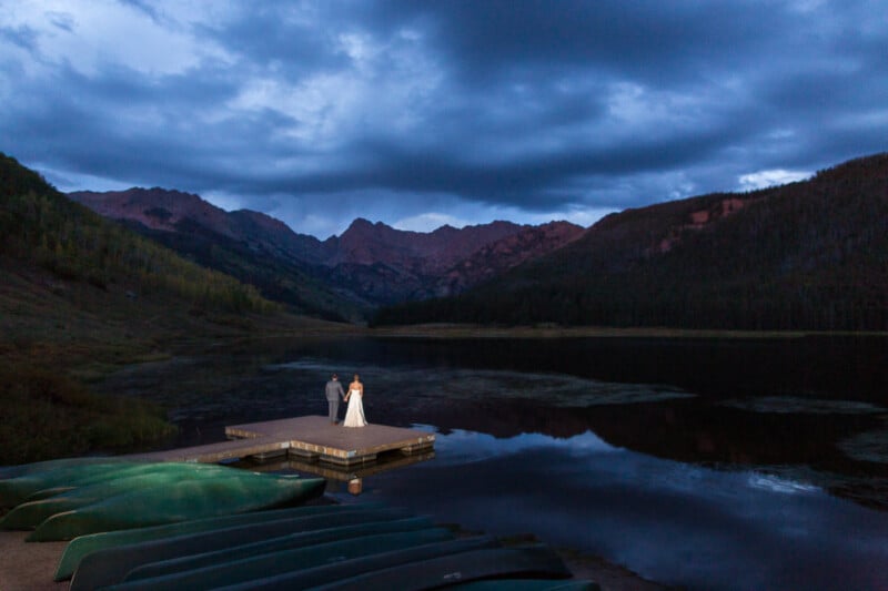 A couple stands on a dock at dusk, overlooking a calm lake surrounded by mountains. The sky is filled with dramatic clouds, and canoes are visible in the foreground.