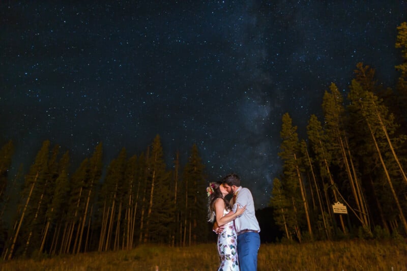 A couple embraces under a starry night sky with the Milky Way visible above. Tall trees line the background, creating a serene and romantic atmosphere.