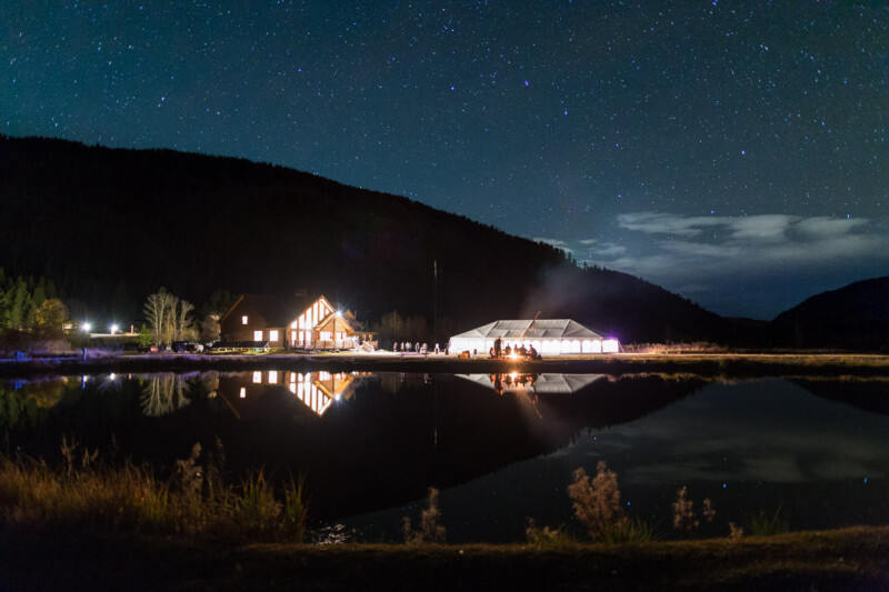 A serene night scene with a waterfront building and a lit tent reflected in the calm water. A group of people gathered around a campfire are visible under a starry sky, surrounded by mountains.