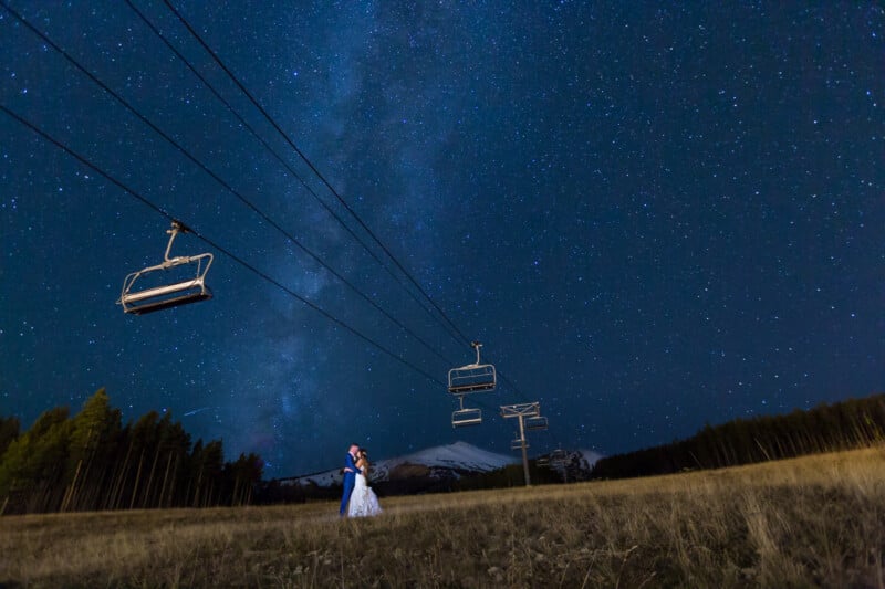 A couple in wedding attire stands on a grassy field under a clear, starry night sky. Empty ski lift chairs hang overhead. A distant mountain is visible, framed by trees on either side.