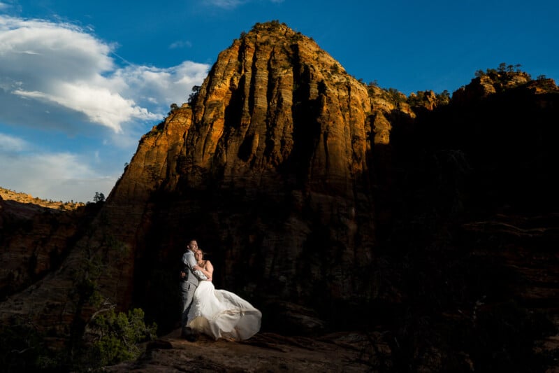 A couple in wedding attire embrace on a rocky outcrop at sunset. The bride's dress flows elegantly, while a towering cliff bathed in golden light looms behind them, contrasting against a bright blue sky.