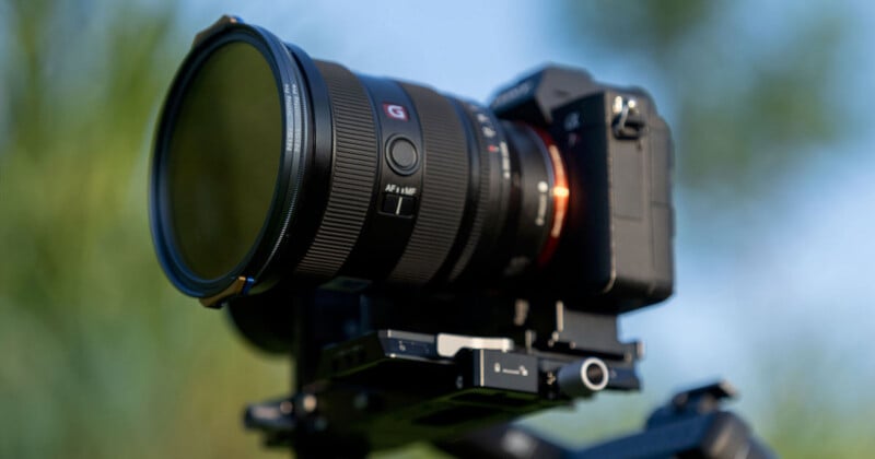 A close-up view of a professional camera mounted on a tripod. The camera features a large lens with branding visible, set against a blurred natural background.